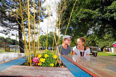 Dos personas disfrutan bebidas en una mesa con flores en el camping Seasonova Camping Le Brabois en Grand Est, Francia.