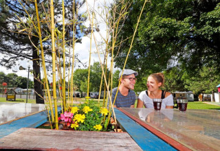 Dos personas disfrutan bebidas en una mesa con flores en el camping Seasonova Camping Le Brabois en Grand Est, Francia.
