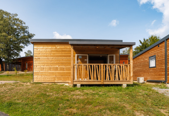 Holzhütte mit Veranda und großen Fenstern, frontal fotografiert unter blauem Himmel mit Wolken.