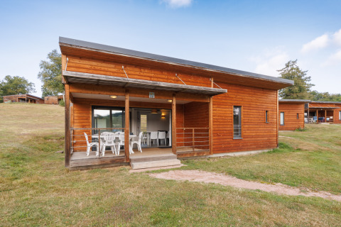 Holzlodge mit überdachter Terrasse und weißen Gartenmöbeln auf einer Wiese, umgeben von Natur.