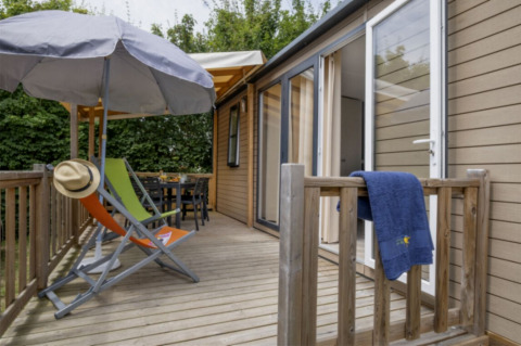 Holzterrasse am Mobile home Confort mit Liegestühlen, Sonnenschirm, Hut, Handtuch und Gartenblick.