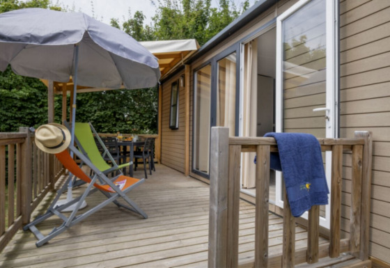 Wooden deck at Mobile home Confort featuring sun loungers, umbrella, hat, towel and garden view.