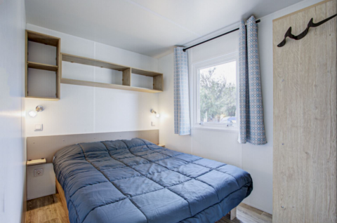 Premium Mobile Home soveværelse med dobbeltseng, reoler, vindue og lysgardiner på Seasonova Camping Barberousse.