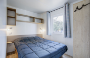 Slaapkamer van Premium Mobile Home bij Seasonova Camping Barberousse met tweepersoonsbed, planken en blauwe gordijnen.