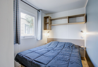 Slaapkamer met dubbel bed, blauw dekbed, wandplanken en raam in Premium Mobile Home in Frankrijk.