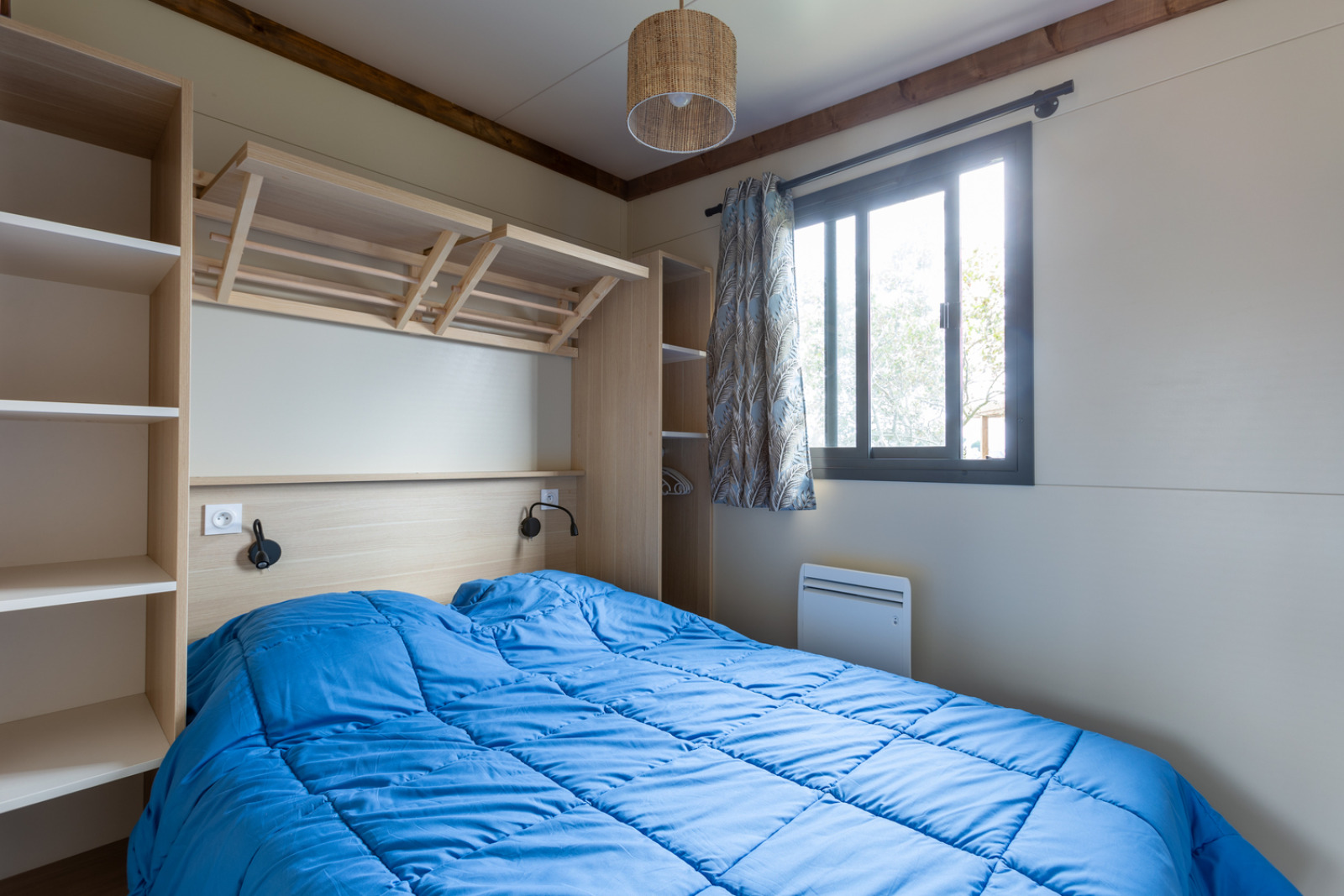 Slaapkamer met tweepersoonsbed, blauw dekbed, planken en raam in Cottage Premium bij Camping Seasonova La Pointe de Roscoff.