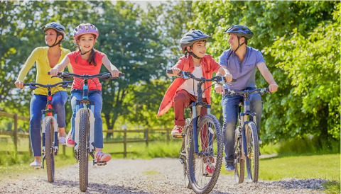 Une famille fait du vélo ensemble sur un chemin de gravier, ensoleillé, entouré d’arbres et de verdure.