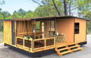 Casa diminuta moderna en el bosque con porche de madera, área de estar exterior y rodeada de árboles altos.