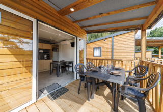 Covered wooden lodge terrace with outdoor dining table set, open kitchen in the background, and chairs.