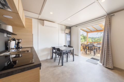 Moderne lodge met keuken, eethoek en overdekt terras met plastic stoelen en uitzicht op groen landschap.