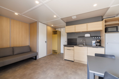 Modern lodge room interior with kitchenette, sofa, dining table, fridge, and light wooden accents.