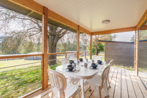 Überdachte Terrasse einer Lodge mit weißem Tisch, sechs Stühlen und ruhiger Aussicht ins Grüne.