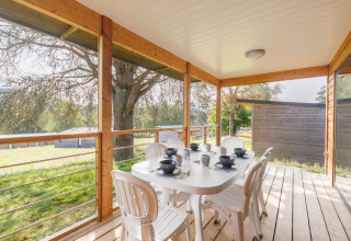 Überdachte Terrasse einer Lodge mit weißem Tisch, sechs Stühlen und ruhiger Aussicht ins Grüne.