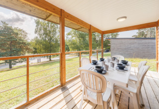 Terrasse abritée d’un lodge avec table dressée, chaises blanches, vue sur pelouse et arbres verdoyants.