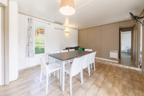 Luminoso comedor moderno en una cabaña con seis sillas blancas, mesa grande y suelo de madera clara.