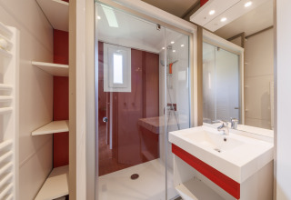 Baño moderno en un lodge con ducha de vidrio, lavabo blanco con detalles rojos y estantes integrados.