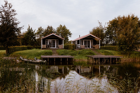 To hyggelige lodgehytter ved en sø omgivet af grønt, med små både ved bådebroerne foran husene.