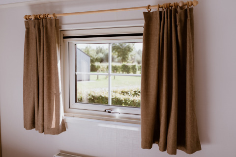 Window with brown curtains inside Chalet lodge at Holiday park Eigen Wijze, Netherlands, overlooking garden.