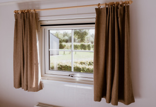 Ventana con cortinas marrones en Chalet lodge en Holiday park Eigen Wijze, Países Bajos, con vista al jardín.