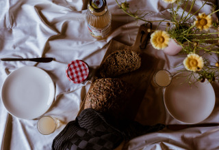 Frühstückstisch in einer Lodge mit Brot, Saft, Marmelade, zwei Tellern und gelben Blumen.
