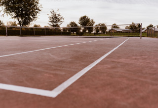 Gros plan sur un court de tennis vide dans un lodge, entouré d'arbres, de clôtures et d’un ciel nuageux.