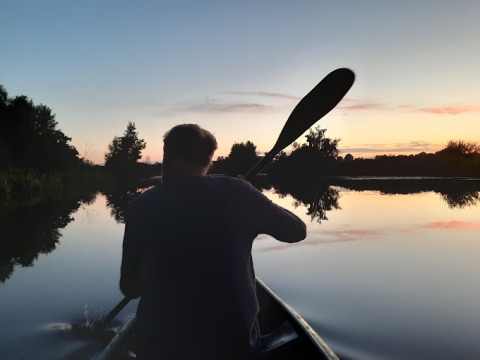 Person paddling a canoe on a calm lake at sunset, surrounded by trees, near a lodge retreat area.
