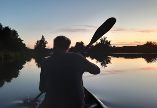 Person padler i en kano på en rolig sø ved solnedgang, omgivet af træer, set fra et lodgeområde.