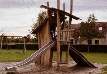 Speelplein met houten klimrek en glijbaan aan een glampinglocatie, omringd door gras en huizen.