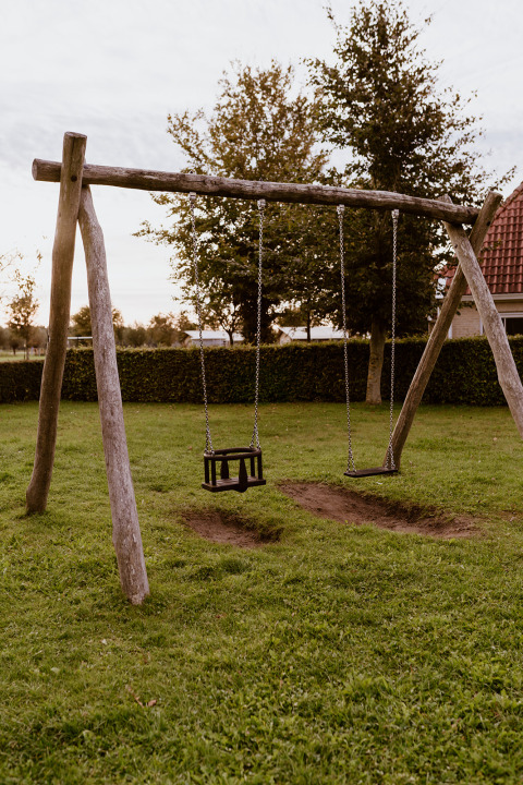 Holzschaukel auf einer grünen Wiese bei einer Glamping-Unterkunft. Bäume stehen im Hintergrund.