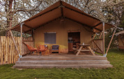 Safarizelt mit Holzveranda, orange Stühlen, Picknicktisch und blühenden Bäumen im Hintergrund.