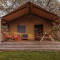 Safaritent met houten veranda, oranje stoelen, picknicktafel en bloesemende bomen op de achtergrond.