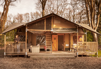 Safari tent De Bosvilla with stove, set in a wooded area, two people standing on the front porch.