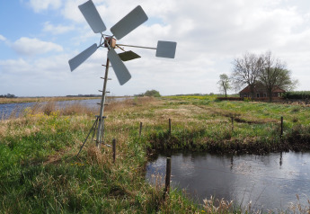 Lille hus og vindmølle ved vand på feriestedet Eigen Wijze i Holland, omgivet af grøn natur.