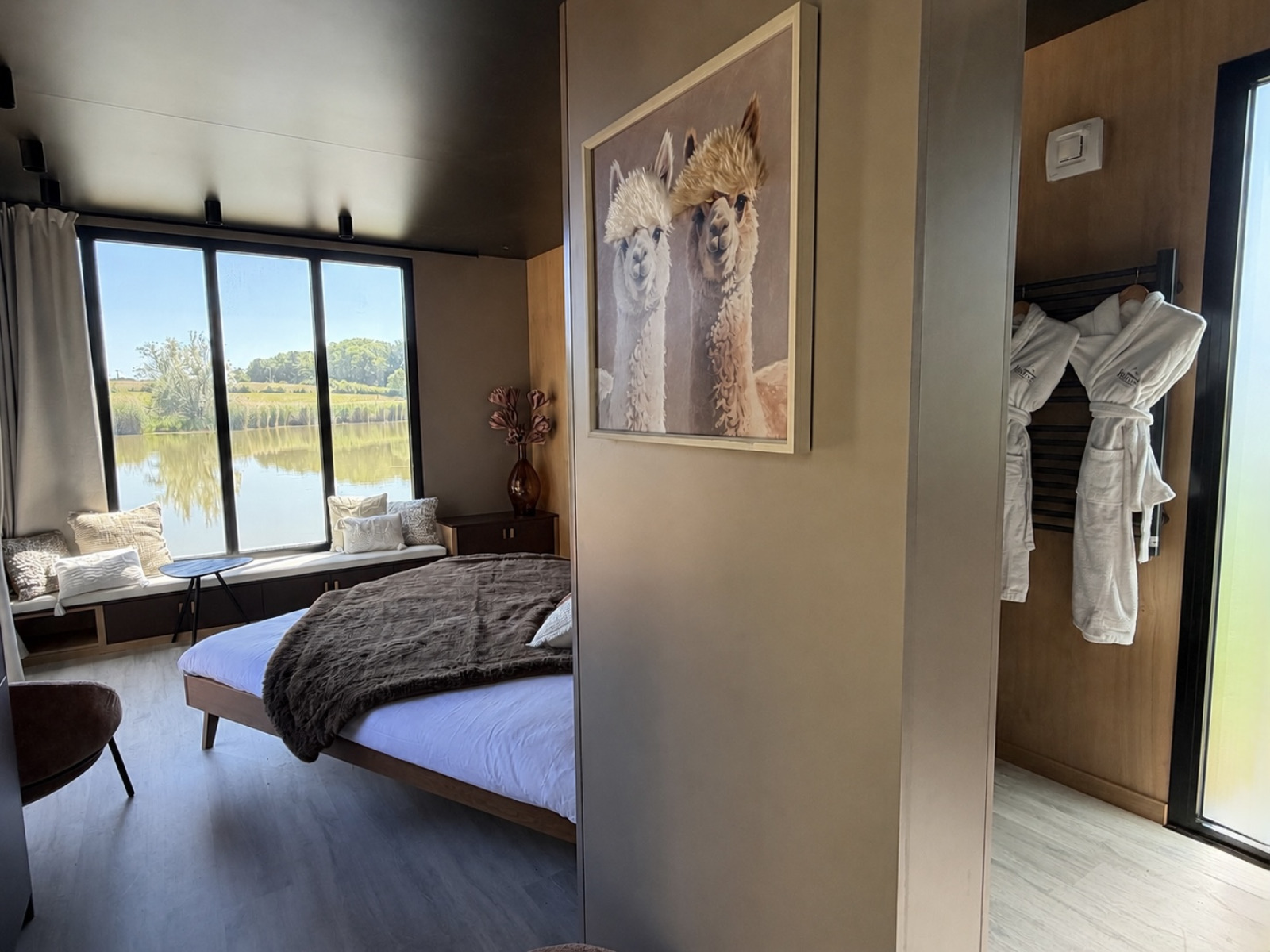 Moderne lodgekamer met uitzicht op het meer, knus bed, grote ramen en een alpaca-kunstwerk aan de muur.