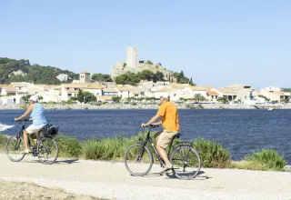 To cyklister kører langs vandet med udsigt til Gruissan og et gammelt tårn i Occitanie, Frankrig.
