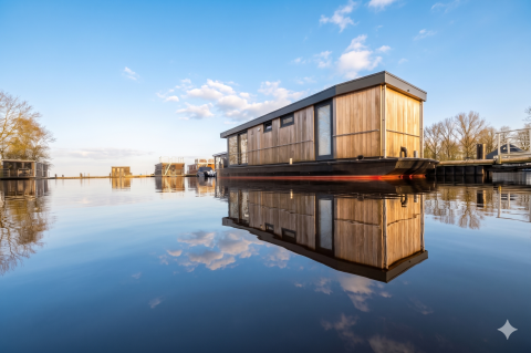 Modern wooden houseboat reflected on calm water at Welcome In - Friesland, Netherlands, with blue sky.