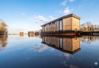 En moderne husbåd lavet af træ flyder på stille vand med blå himmel ved Welcome In - Friesland, Nederlandene.
