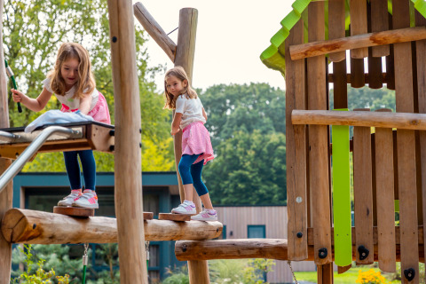 Two girls playing on a wooden playground at a lodge, with trees and modern cabins in the background.