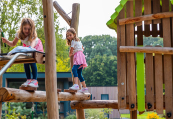 Twee meisjes spelen op een houten speeltoestel bij een lodge, met bomen en lodges op de achtergrond.