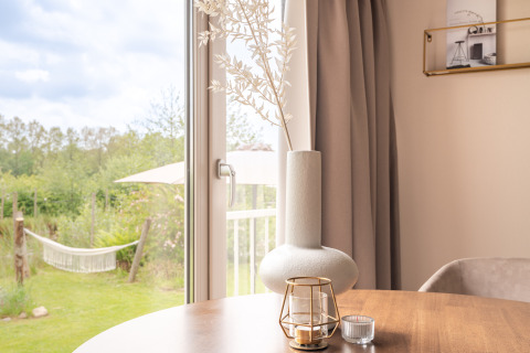 Licht interieur met vaas en kaars op tafel, uitzicht op tuin en hangmat door glazen deur.
