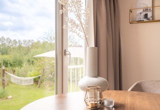 Licht interieur met vaas en kaars op tafel, uitzicht op tuin en hangmat door glazen deur.