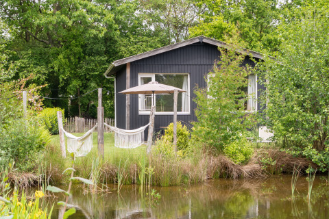 Zwart tiny house bij een vijver, omgeven door groen, met hangmatten en een parasol, De Kremmer, Nederland.
