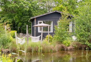 Black tiny house by a pond surrounded by greenery, with hammocks and an umbrella, De Kremmer, Netherlands.