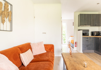 Salon lumineux de tiny house avec canapé orange, table en bois, cuisine moderne et chambre en arrière-plan.