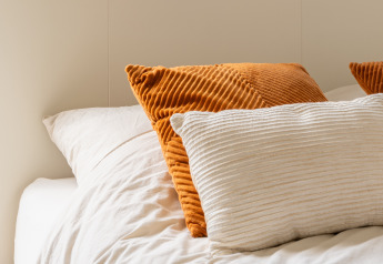 Gros plan d’un lit douillet en glamping avec draps blancs et coussins orange et crème.