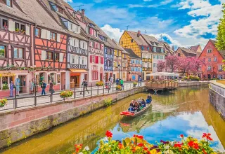 Farverig kanal med bindingsværkshuse og blomster i Bassemberg, Grand Est, Frankrig, med folk i båd.