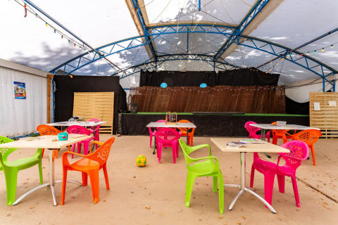 Farverig udendørs café med plastikstole og borde, scene med lyskæder på Camping Paradis Le Giessen.