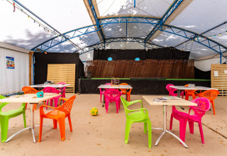 Sillas y mesas de plástico coloridas en una carpa exterior con escenario en Camping Paradis Le Giessen, Francia.