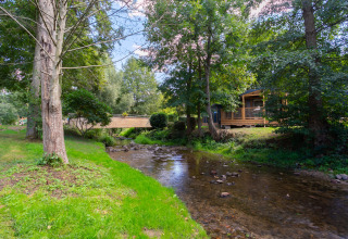 En fredelig campingplads ved en bæk med grønne træer og en hytte, set i Camping Paradis Le Giessen, Grand Est.