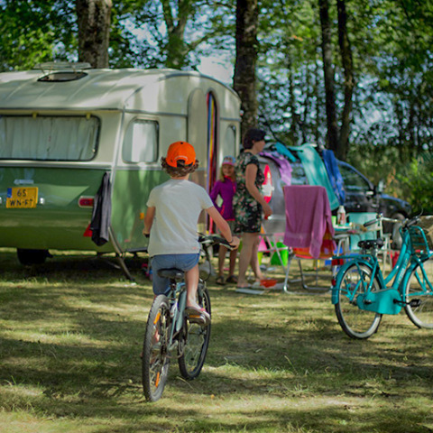Kinder fahren Fahrrad an einem Wohnwagen im Schatten, während Familien in einem französischen Ferienpark entspannen.